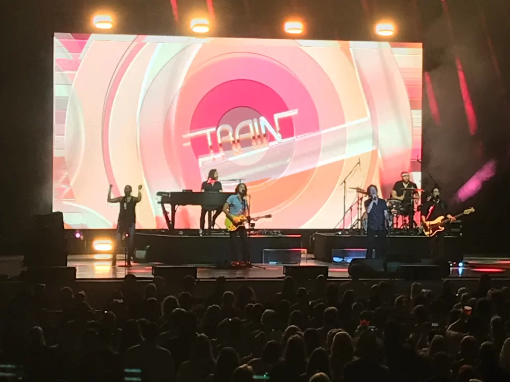 Train band engaging with fans during a live performance at The Orion Amphitheater, featuring weather contingency measures